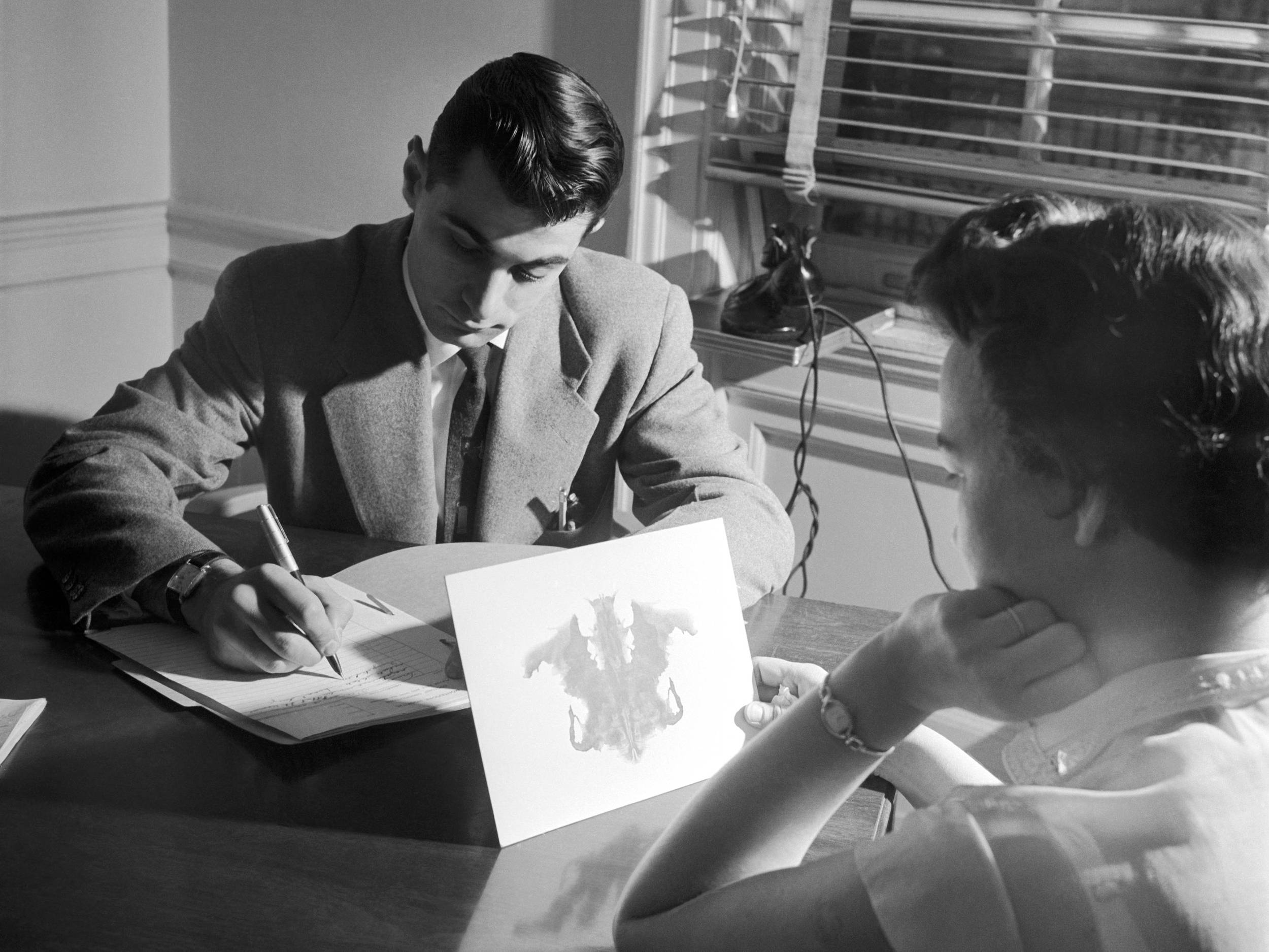 Fotografie, schwarz-weiß, um 1950: Das Foto zeigt einen Mann und eine Frau an einem Tisch sitzend. Der Mann schreibt, sein Blick ist gesenkt. Die Frau betrachtet ein Tinten-Kleks-Bild
