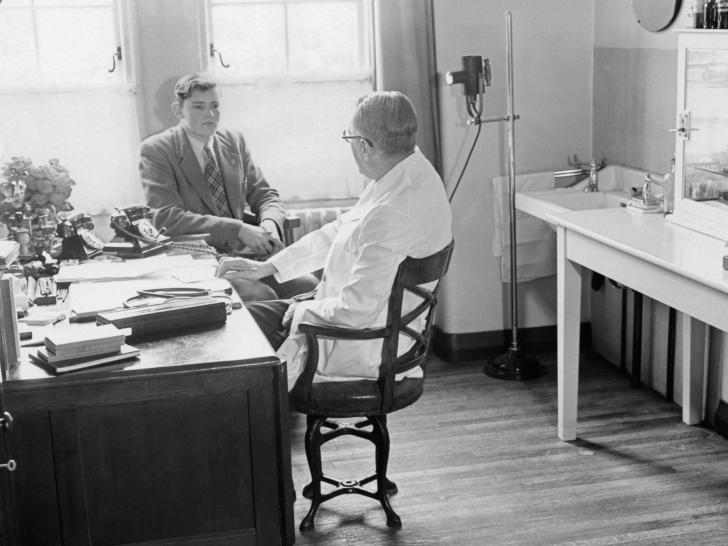 Fotografie, schwarz-weiß, von 1955: Das Foto zeigt einen Patienten und einen Arzt in einem Behandlungszimmer an einem Schreibtisch sitzend. Den Patienten sieht man von vorne. Der Arzt wird in einer Dreiviertelansicht von hinten gezeigt.     