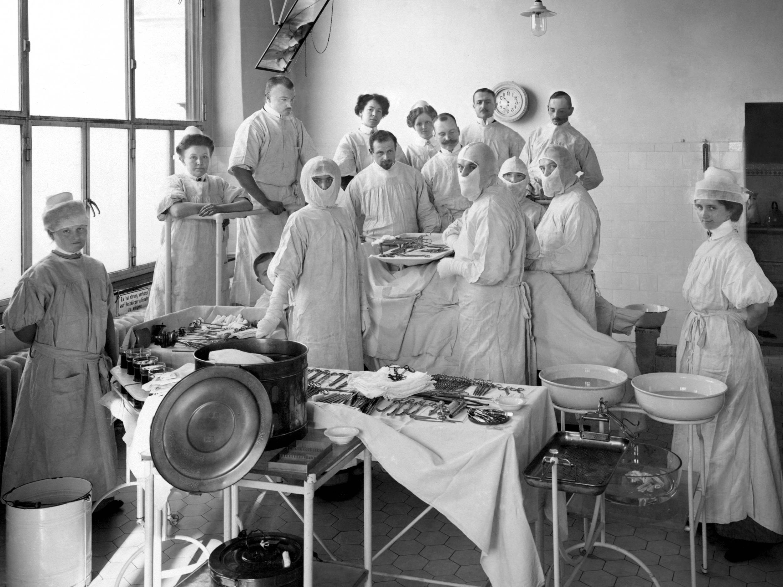 Fotografie, schwarz-weiß, um 1910: Das Foto zeigt fünfzehn Personen in einem Operationssaal. Sie tragen OP-Kleidung, Im Vordergrund liegen OP-Instrumente auf einem Rolltisch, ferner sieht man einen Sterilisator und zwei Emaille-Schüssel mit Wasser.